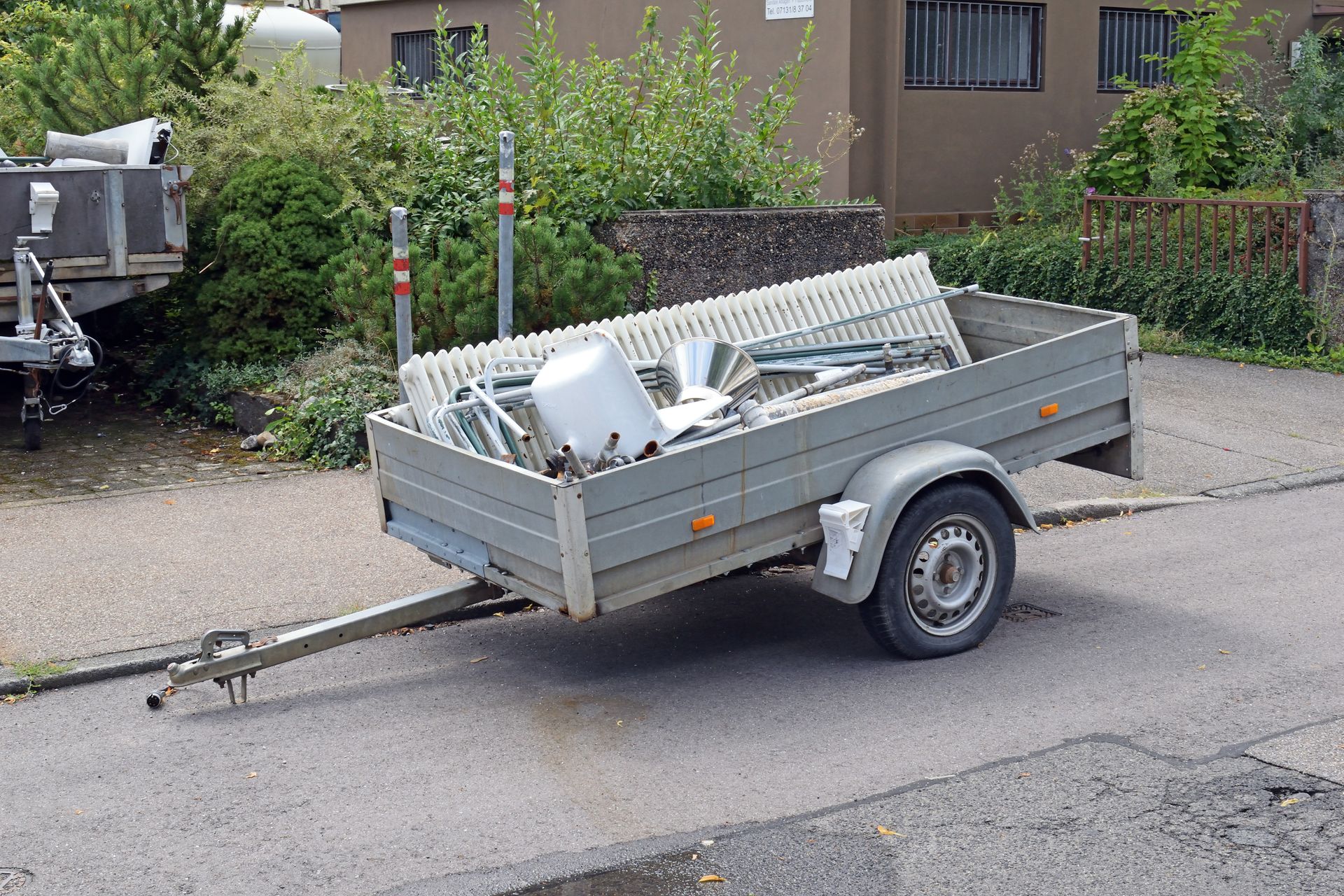 A gray trailer parked on a street, filled with metal scrap. Green bushes and a building in the background.