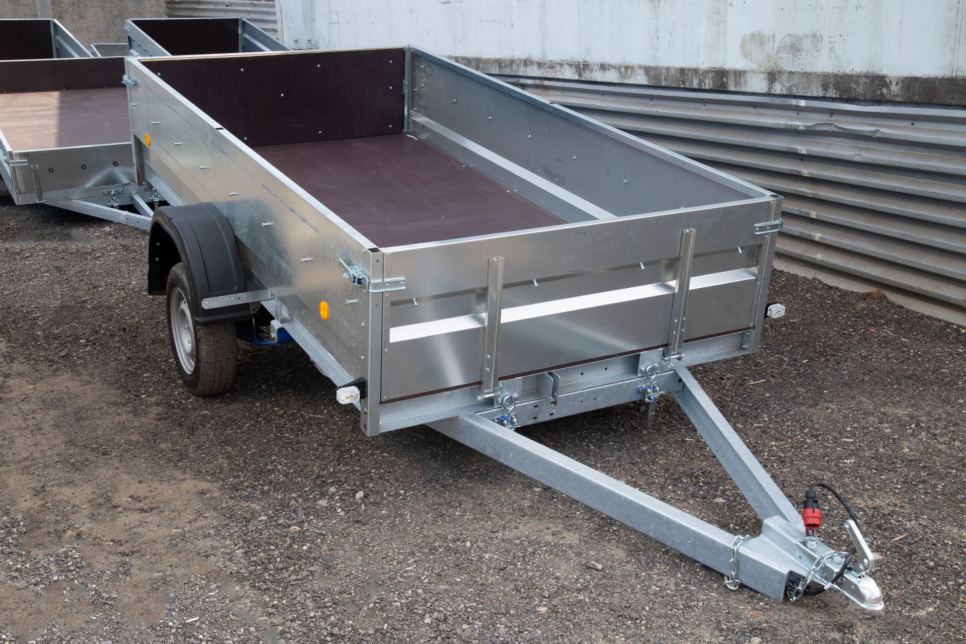 A silver utility trailer with a brown bed sits on gravel, set against a concrete wall and metal siding.