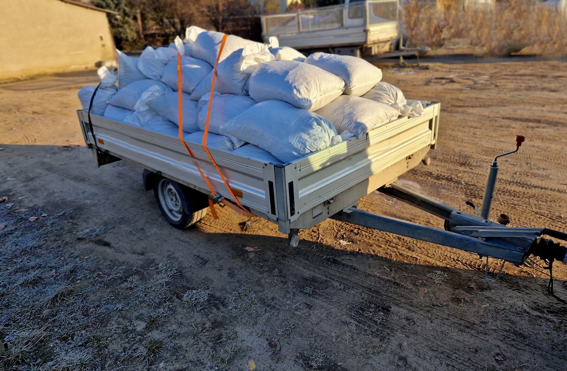 A trailer loaded with numerous white bags, secured by an orange strap, parked on gravel.