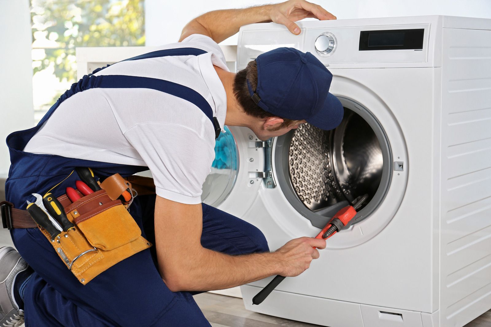 Repairman kneels, working on a washing machine, holding a wrench, and wearing a tool belt.