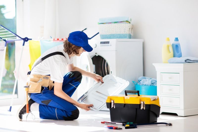 A person in blue overalls and a hat repairs a washing machine in a laundry room.