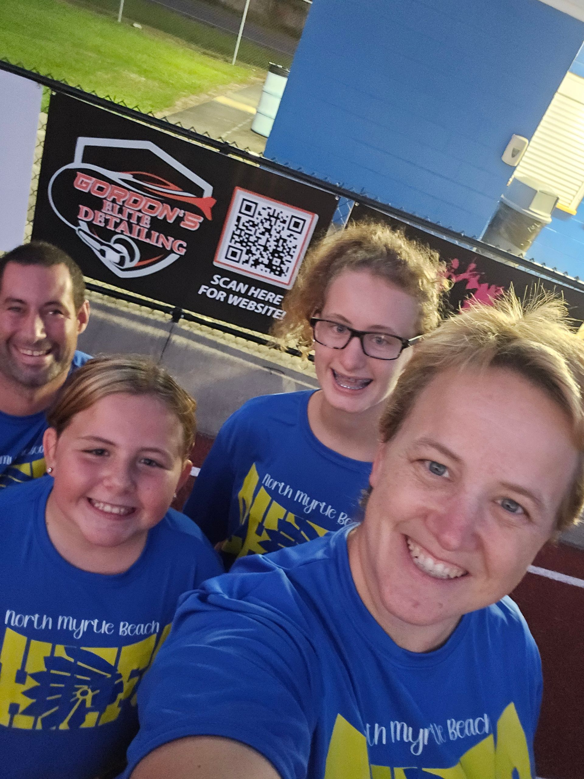 Group of four smiling people in blue shirts; posing for a selfie under a banner at an outdoor event.
