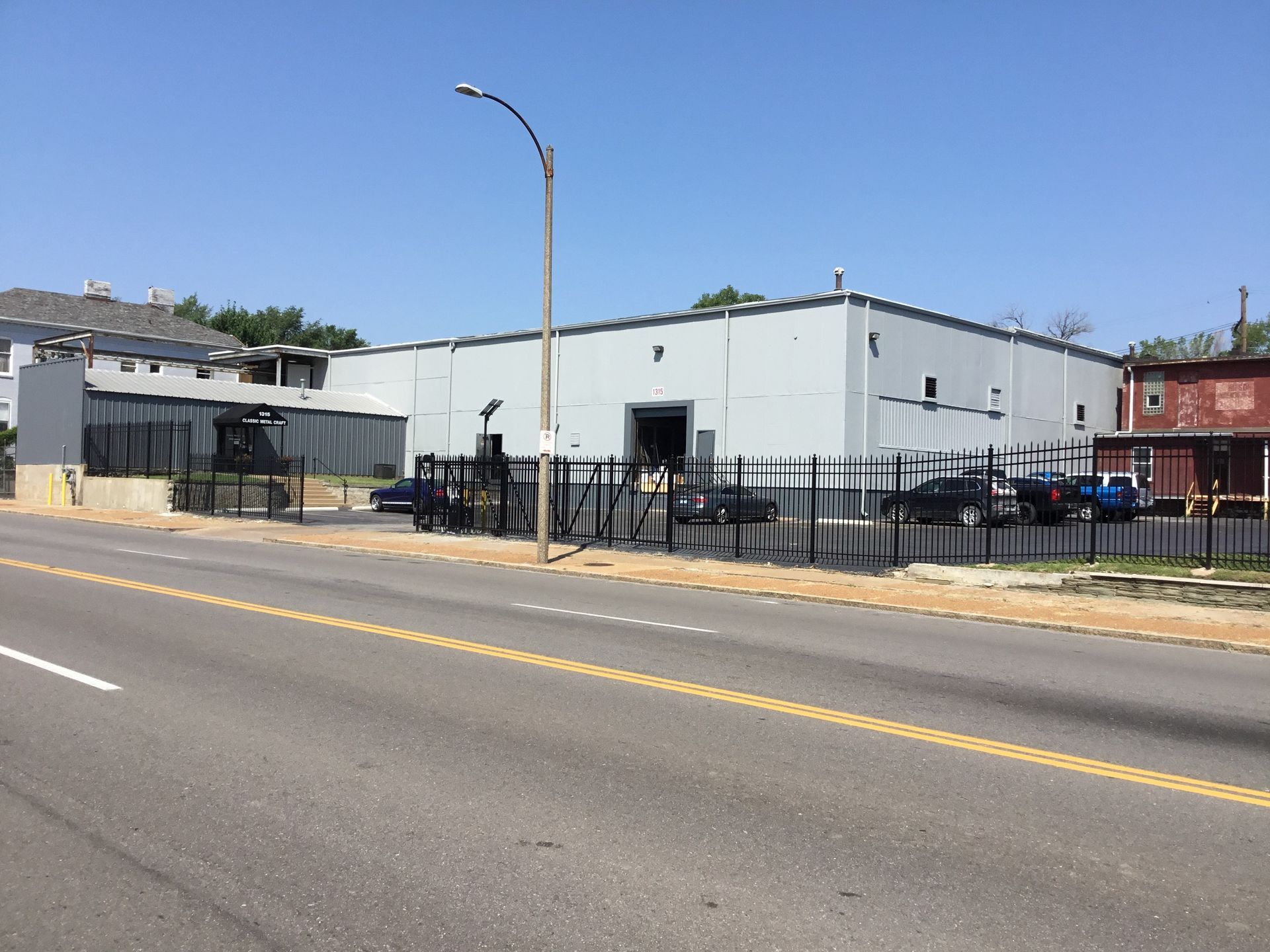 Gray industrial building with black fence along a road on a sunny day.