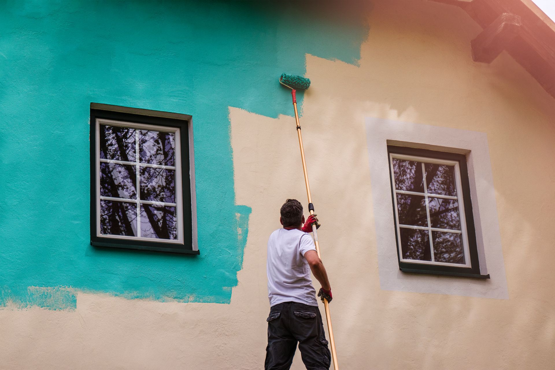 A man is painting the side of a house with a paint roller.