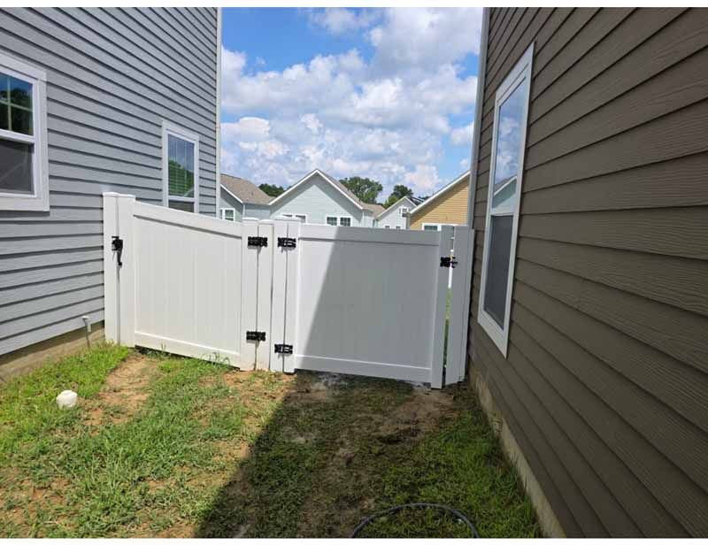 White vinyl gate between two houses along a narrow grassy side yard under a partly cloudy sky