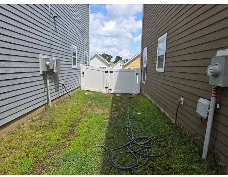 Narrow grassy side yard between two houses with utility meters, a coiled cable, and a white fence gate.