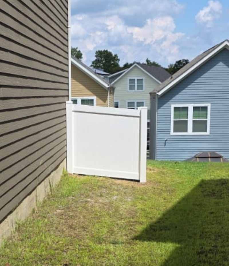 White vinyl gate in a grassy side yard between two houses, with blue and beige siding under a sunny sky.