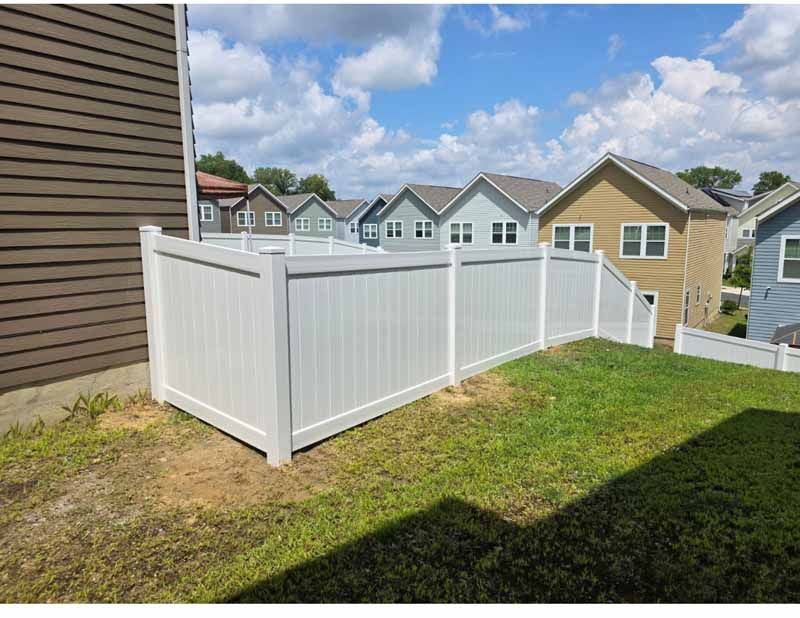 White vinyl fence along a grassy yard beside a house, with suburban homes under a partly cloudy sky.