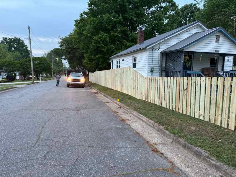 Residential street with a car parked by a white house and wooden fence at dusk