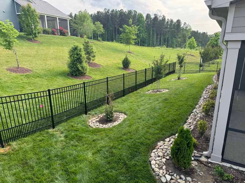 Backyard lawn with black fence, landscaped shrubs, and neighboring houses on a grassy hillside