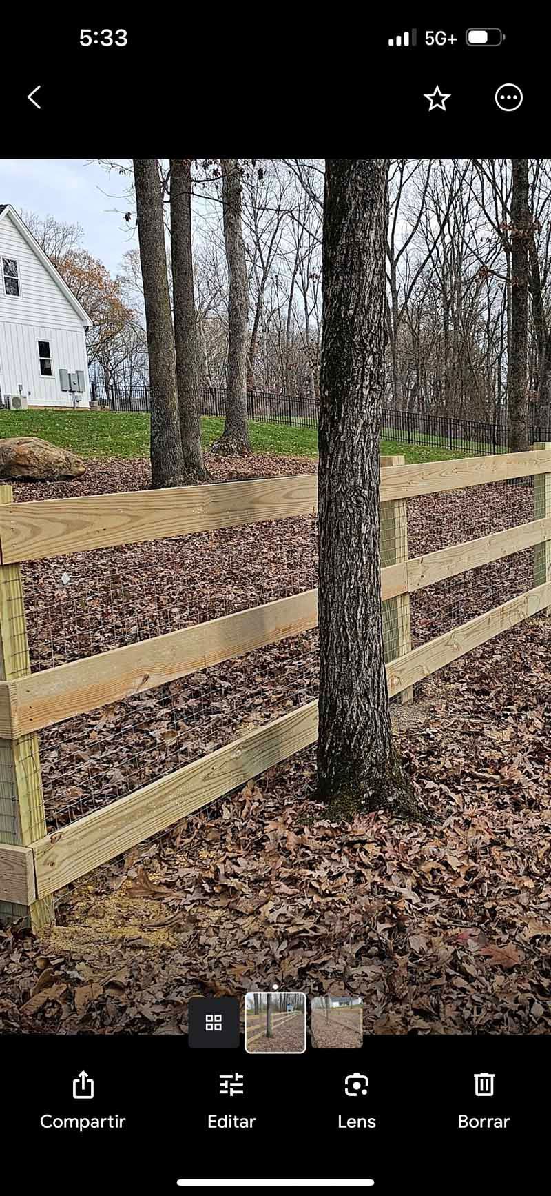 Wooden fence in a leaf-covered yard with bare trees and a white house in the background.