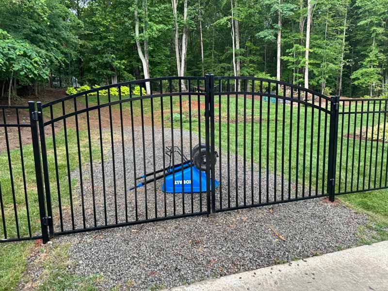 Black metal gate across a gravel path, with a blue bucket or feeder behind it in a wooded yard.