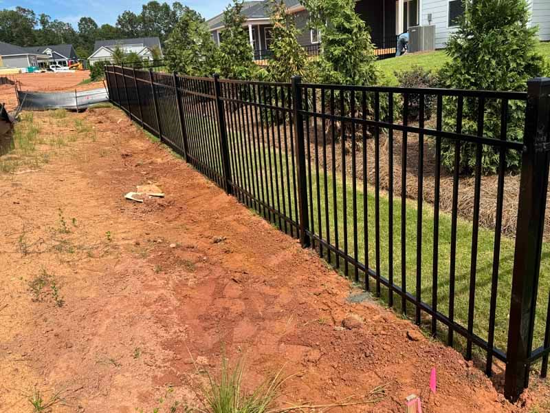 Black metal fence along a red dirt path beside suburban houses and a grassy ditch.