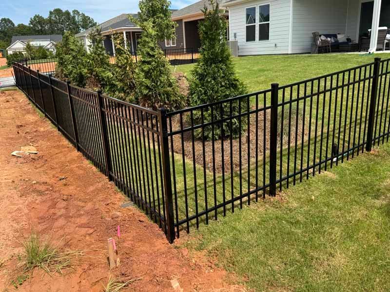 Black metal fence along a grassy yard beside a dirt road in a suburban neighborhood