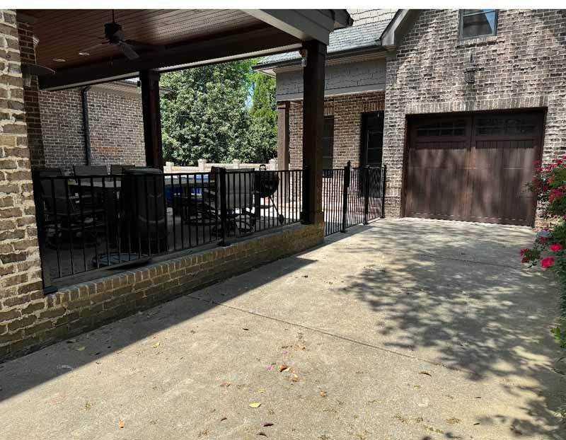 Brick driveway beside a covered patio and garage, with black metal railings and potted flowers in shade