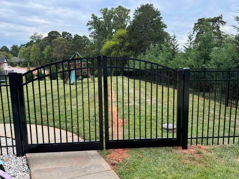 Black metal gate blocking a grassy driveway with trees and houses in the background