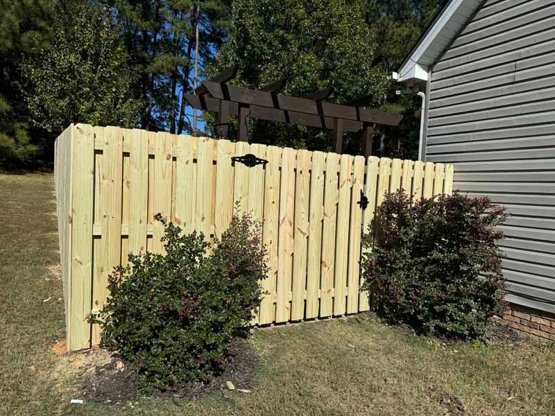 Wooden privacy fence with gate beside a house, bordered by shrubs and trees in a yard