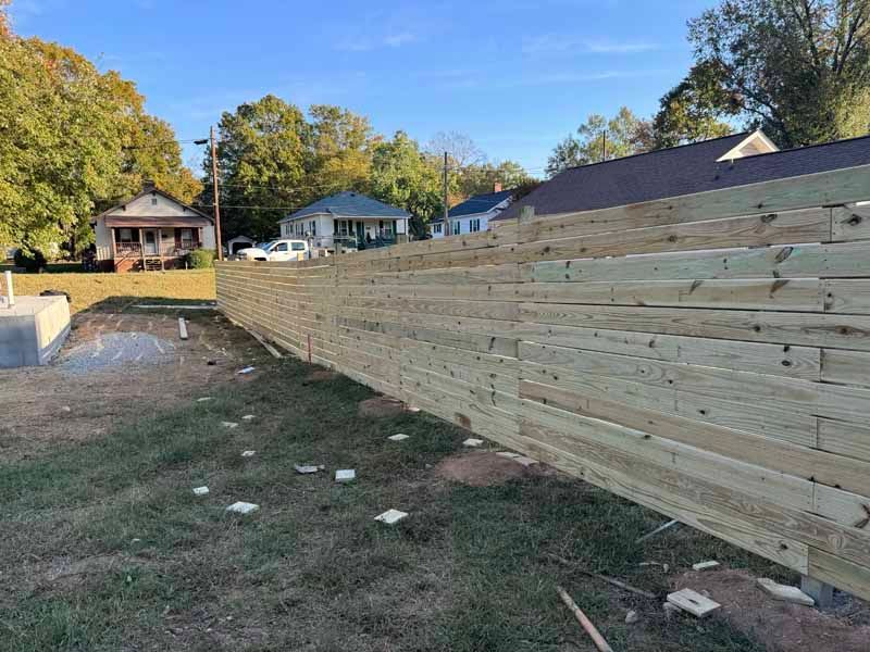 Long wooden retaining wall along a grassy yard under a blue sky with houses in the background