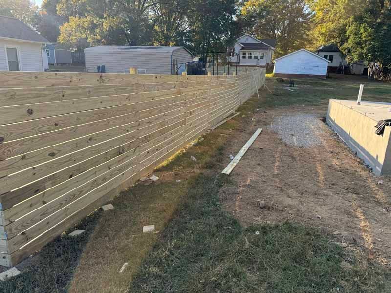 Wooden fence under construction along a backyard path beside a house, with tools and lumber on the ground