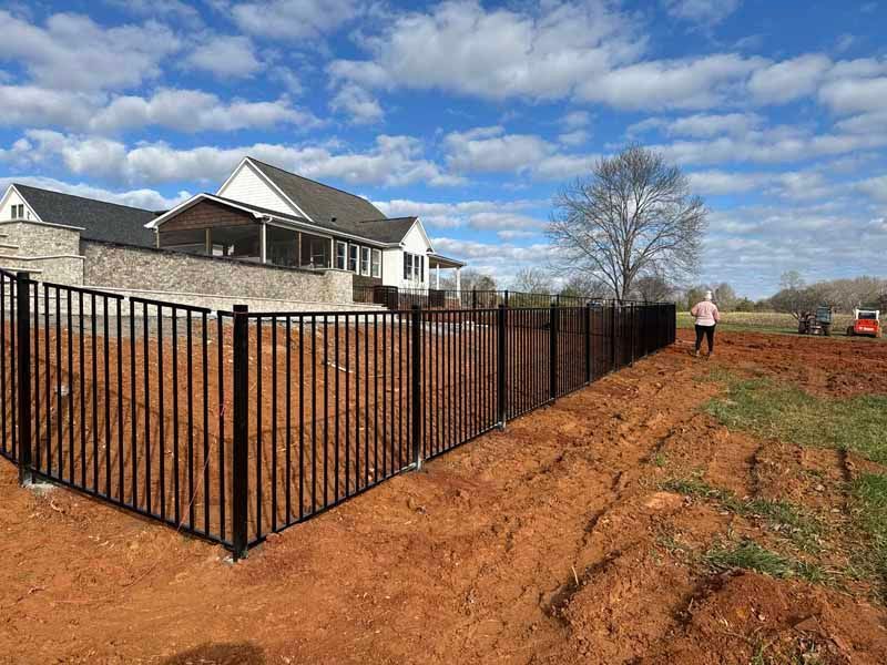 Black metal fence along a house beside a muddy dirt lot under a blue sky