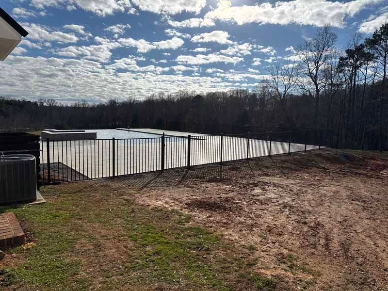 Backyard with white fenced pool, dirt and grass foreground, and cloudy sky over wooded area