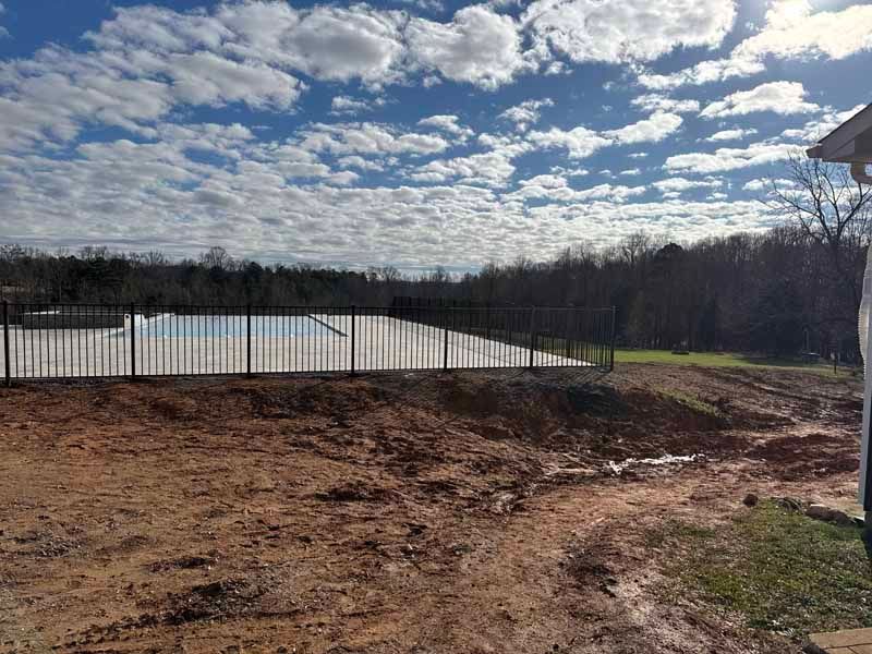 Dirt lot beside a black metal fence under a cloudy sky, with trees in the distance.