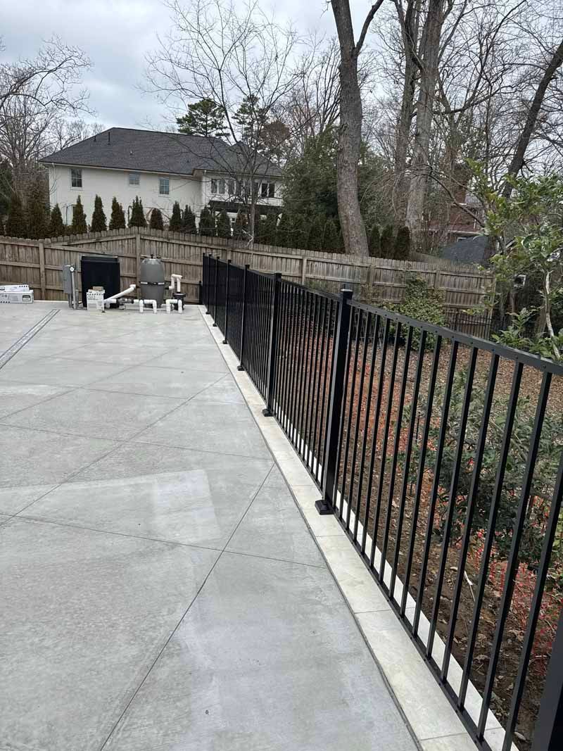 Stone patio with black metal railing overlooking a wooded yard and house beyond