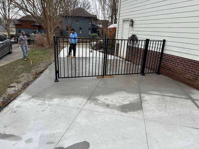 Concrete driveway with a black metal gate blocking access beside a white house; two people stand nearby.