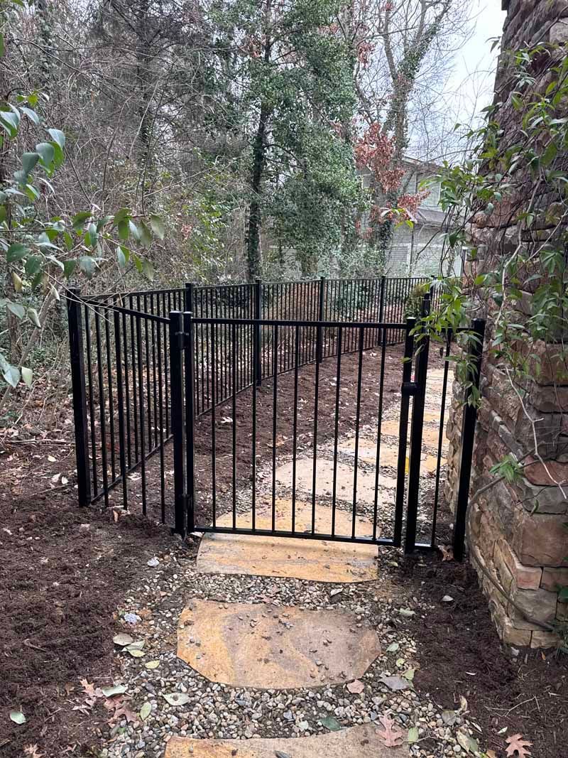 Black metal gate at the end of a stone path, surrounded by trees and a stone wall.