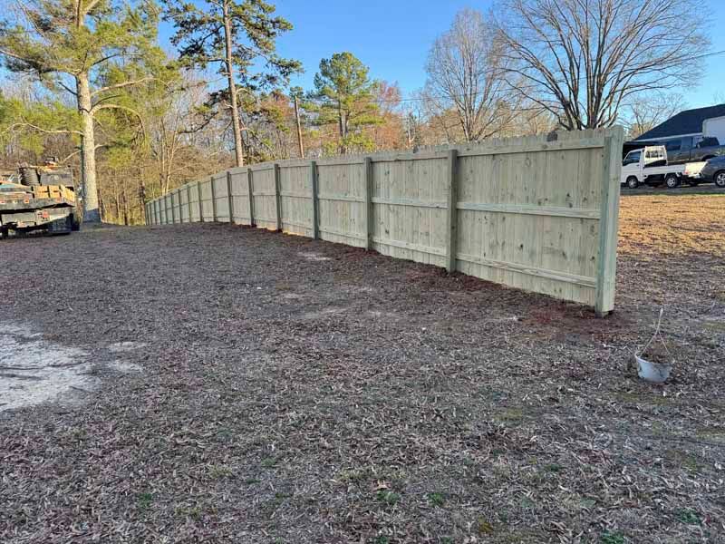 Wooden privacy fence beside a gravel lot with trees and parked vehicles in the background.