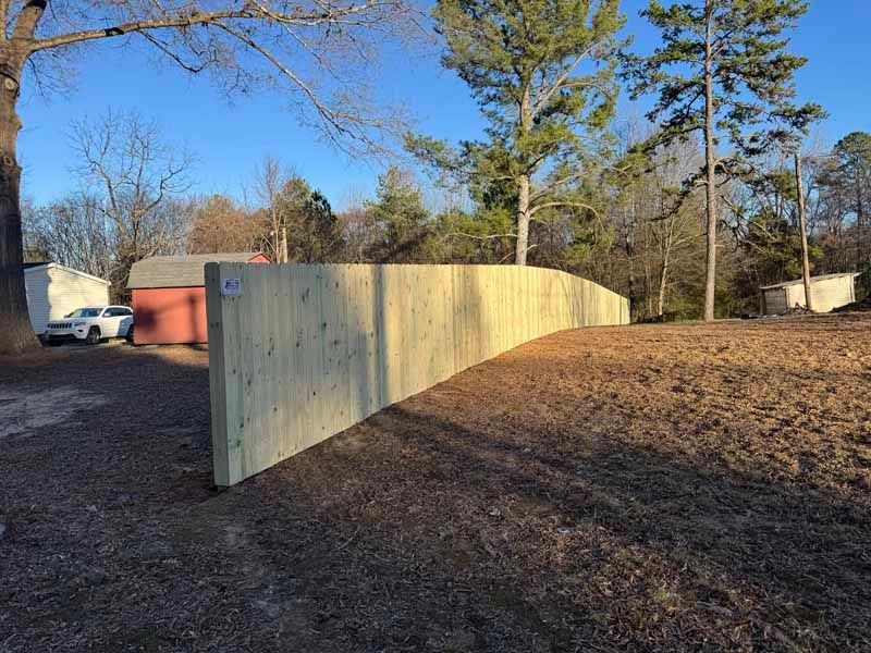 Curved wooden privacy fence in a backyard, with bare trees and a clear blue sky.