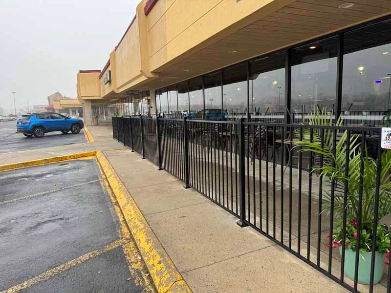 Strip mall sidewalk with black railing, storefront windows, and a blue car in the parking lot