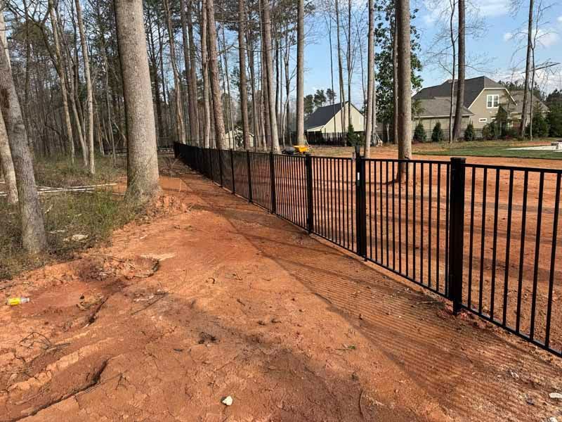 Red dirt path beside a black metal fence in a wooded suburban neighborhood