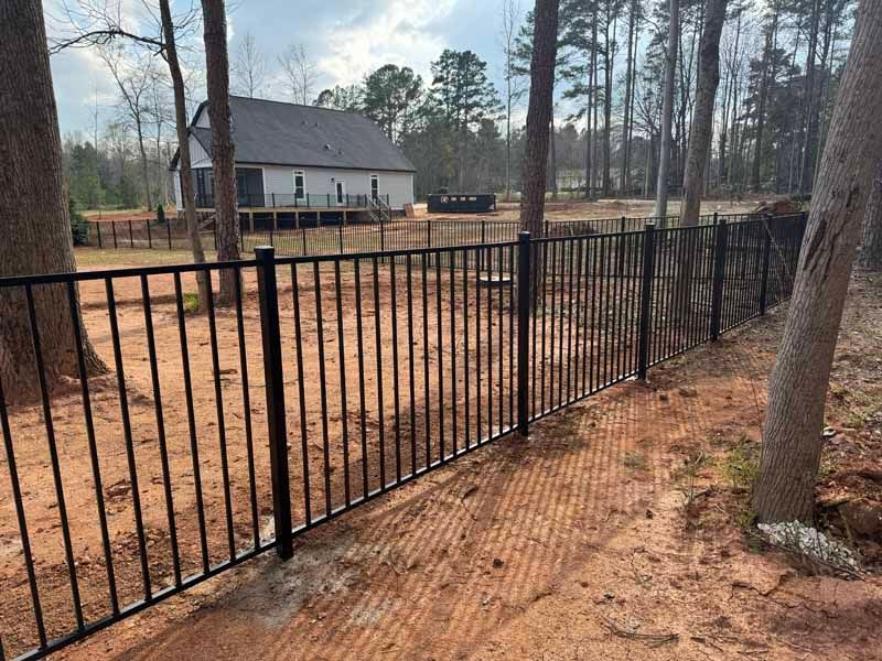 Black metal fence along a dirt yard with a house and tall trees in the background
