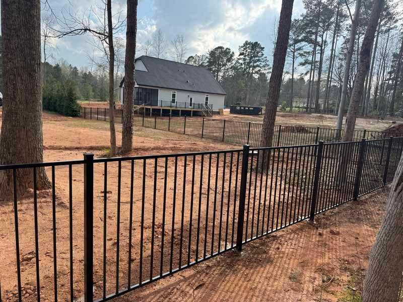 Black metal fence along a dirt yard, with a gray house and trees in the background.