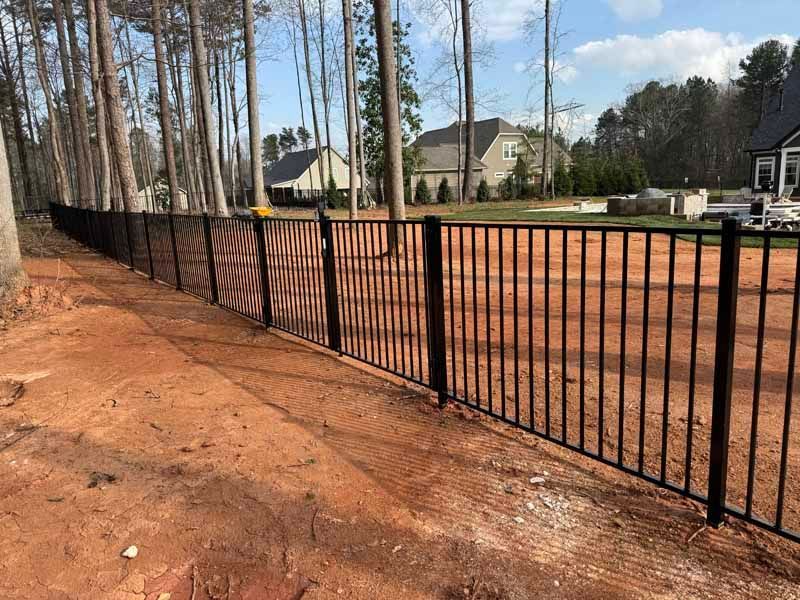 Black metal fence along a red dirt path beside a wooded residential area under a partly cloudy sky