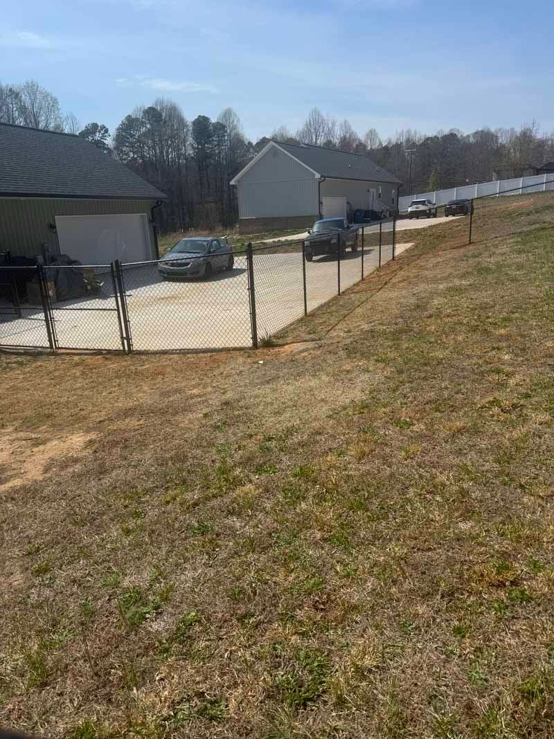Gravel driveway beside a chain-link fence and houses on a sunny hillside.