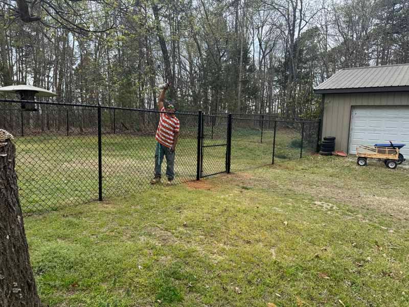 Person installing a black chain-link fence beside a garage in a wooded yard