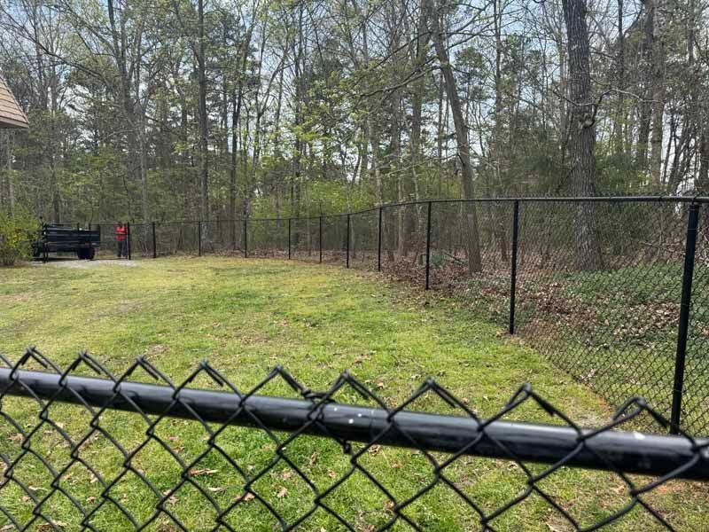 Chain-link fence along a grassy path beside woods, with a person in red near a gate in the distance.