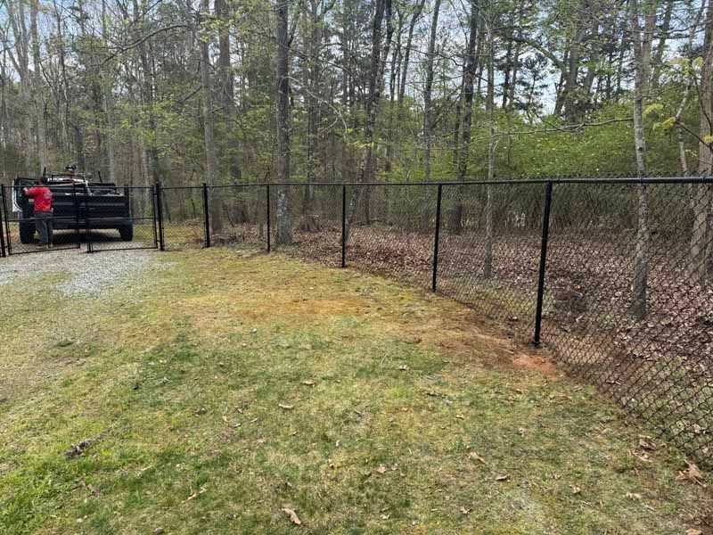 Chain-link fence beside a grassy yard with trees, and a person in a red jacket near a black gate.