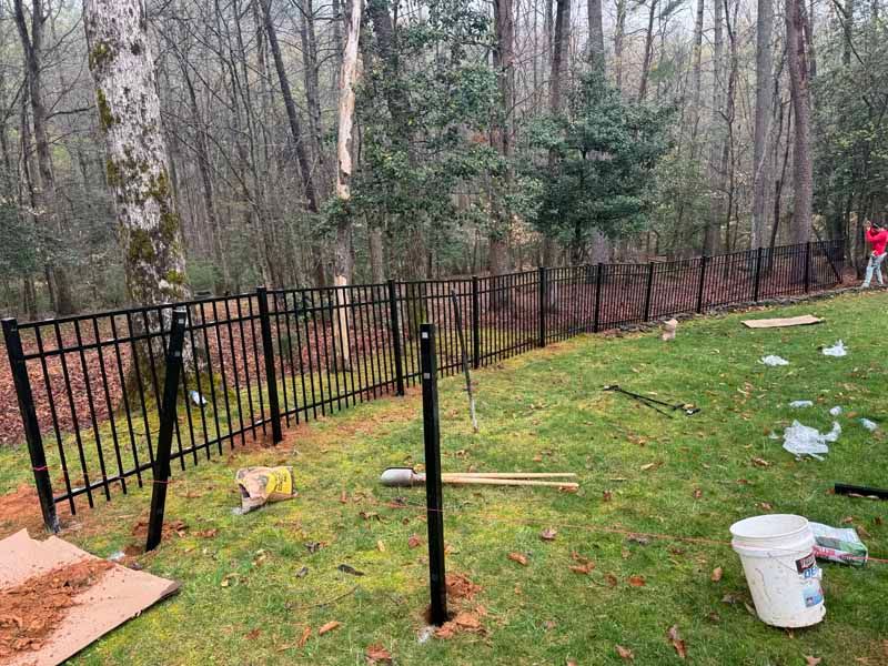Black metal fence beside a grassy yard, with construction debris and a wooded background.