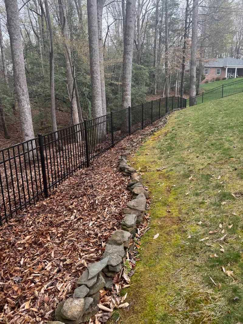 Wooded backyard with a black metal fence, stone border, and grassy slope near a small house