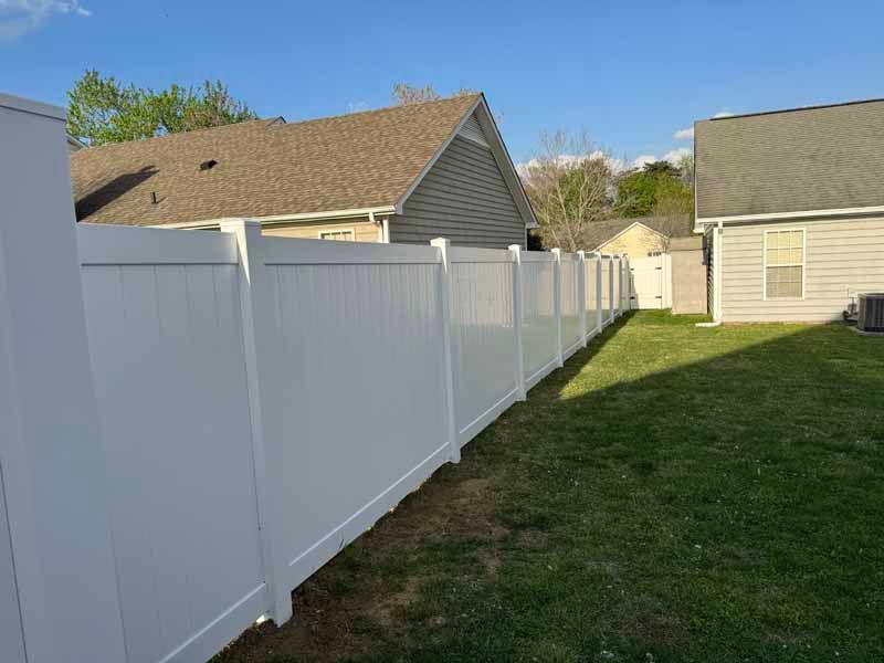 Backyard with a white vinyl fence beside a grassy strip and beige houses under a clear sky