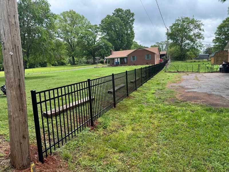 Black metal fence along a grassy yard near a brick house under cloudy skies.