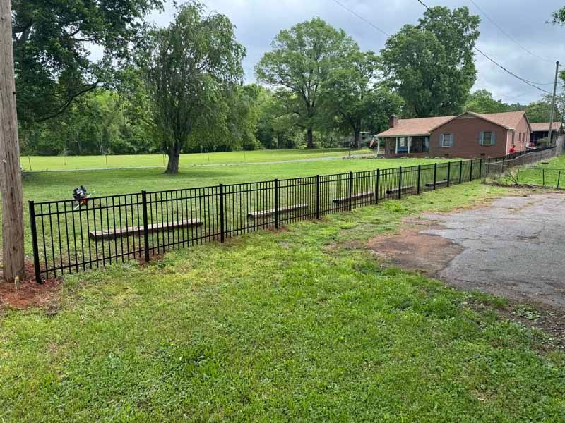 Rustic black metal fence along a grassy yard beside a brick building and paved drive under cloudy skies