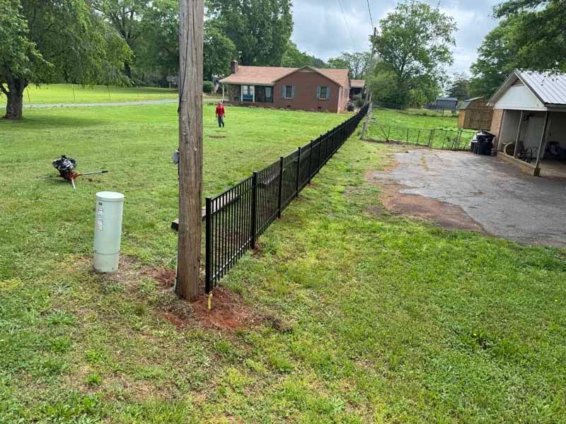 Grassy yard with black fence, utility pole, driveway, shed, and a small brick house in the background.