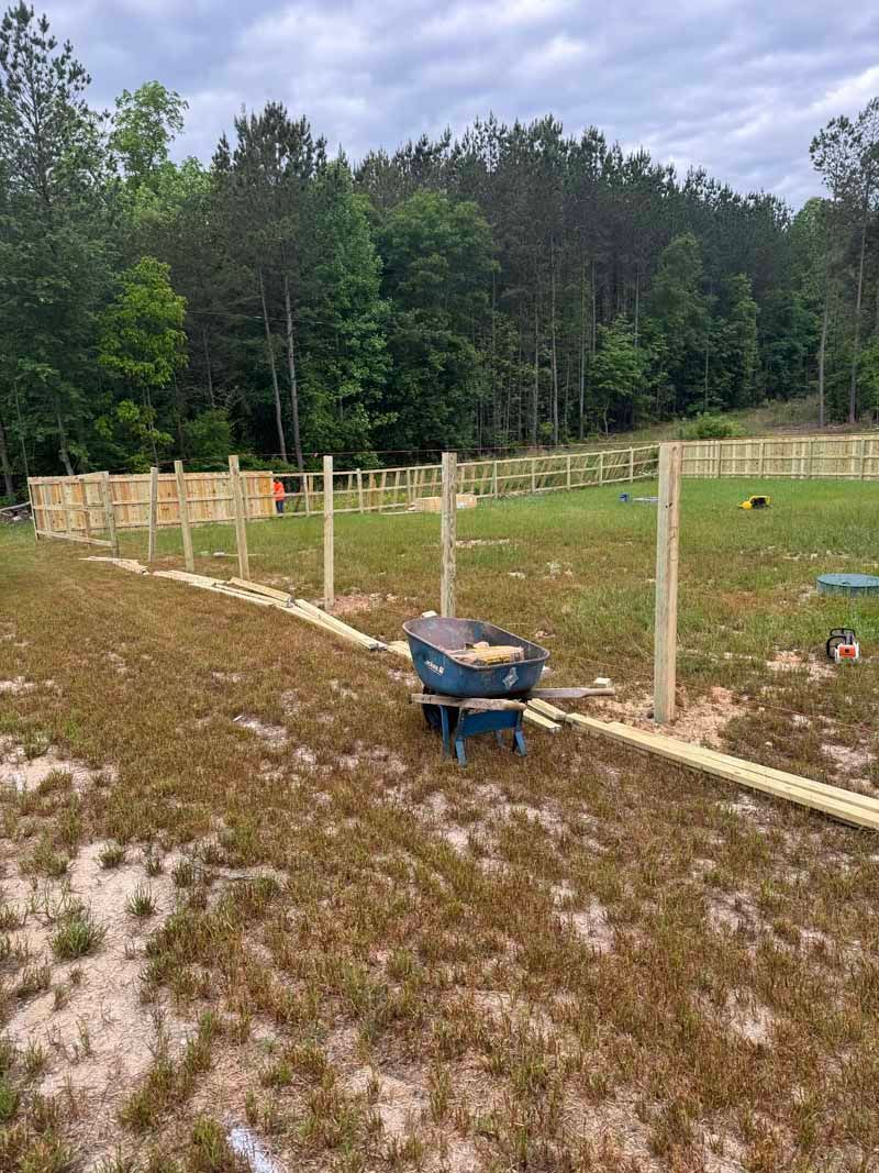 Small fenced grassy field with wooden posts and a blue wheelbarrow beside a dirt path, backed by trees.
