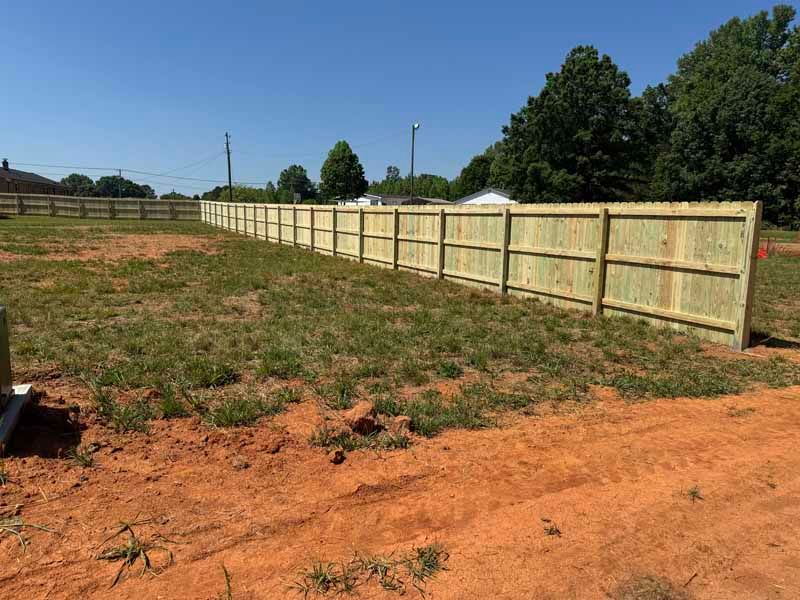Wide dirt lot with a new wooden fence along the right side under a clear blue sky.