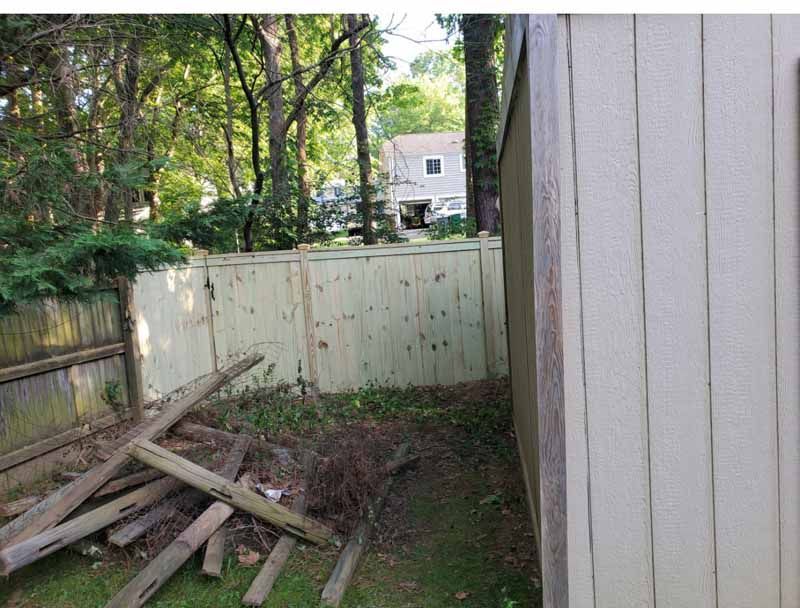 Backyard corner with a white fence, scattered boards, and trees beyond the fence