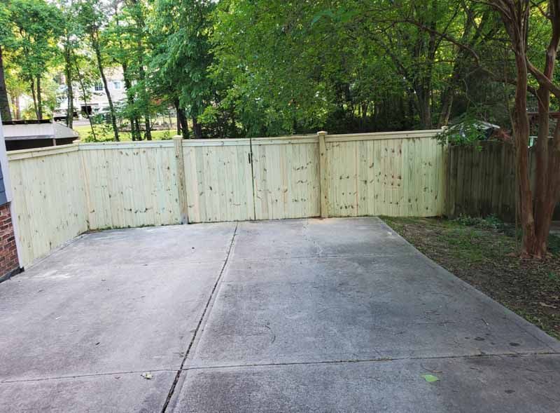 Concrete driveway bordered by a white fence and trees in a wooded backyard
